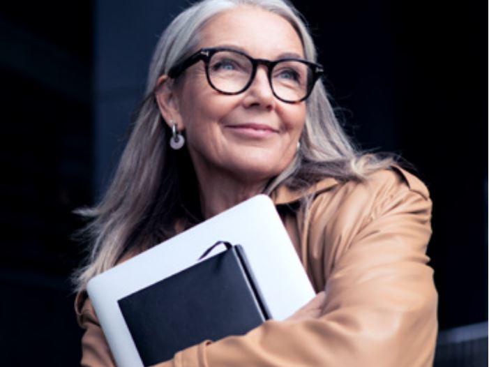 Una imagen de una mujer mayor con lentes que sonríe y lleva un portátil.