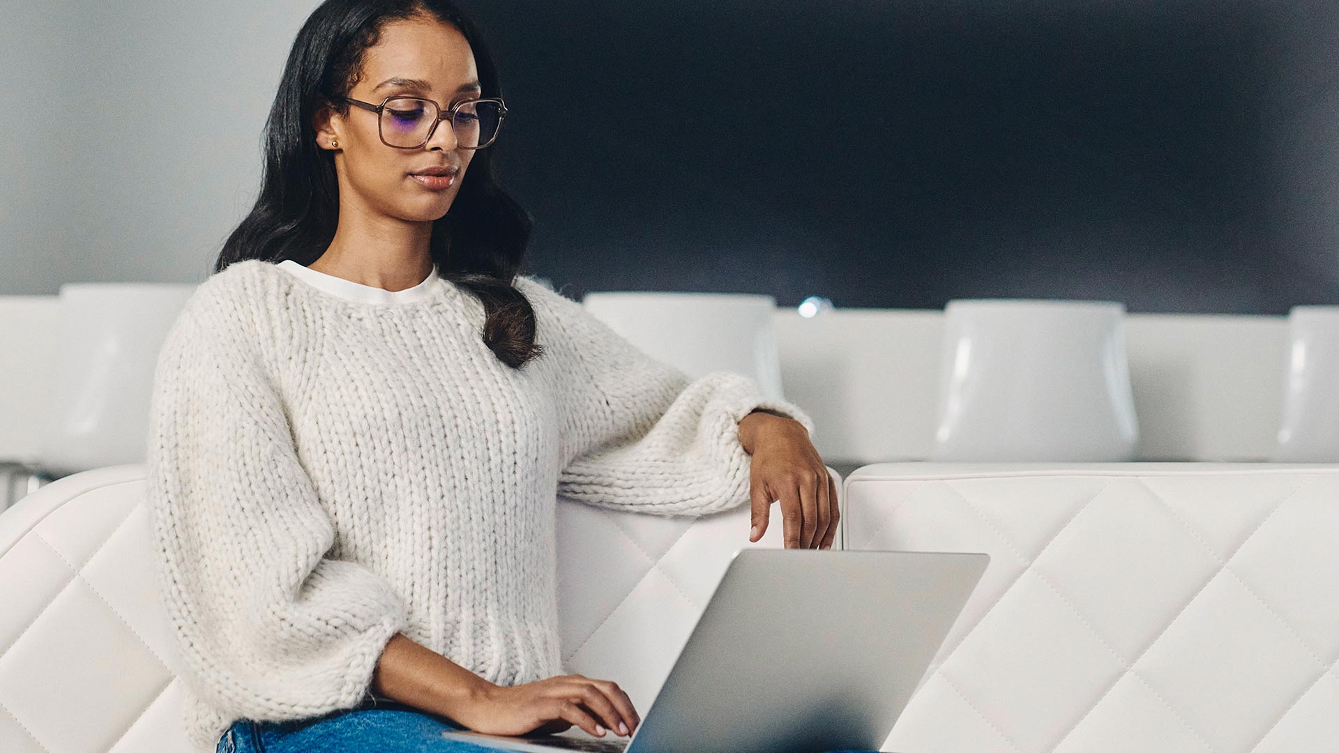Mujer de cabello oscuro, gafas oscuras y suéter blanco utiliza un computador portátil.