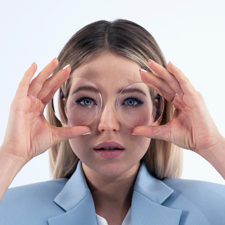 Una joven rubia sostiene unos lentes delante de los ojos para mostrar el efecto de miniojos causado por unos lentes gruesas.