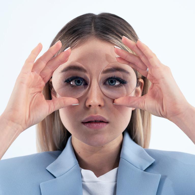 Una joven rubia sostiene unos lentes delante de los ojos para mostrar el efecto de ojo de pez causado por unos lentes gruesas.