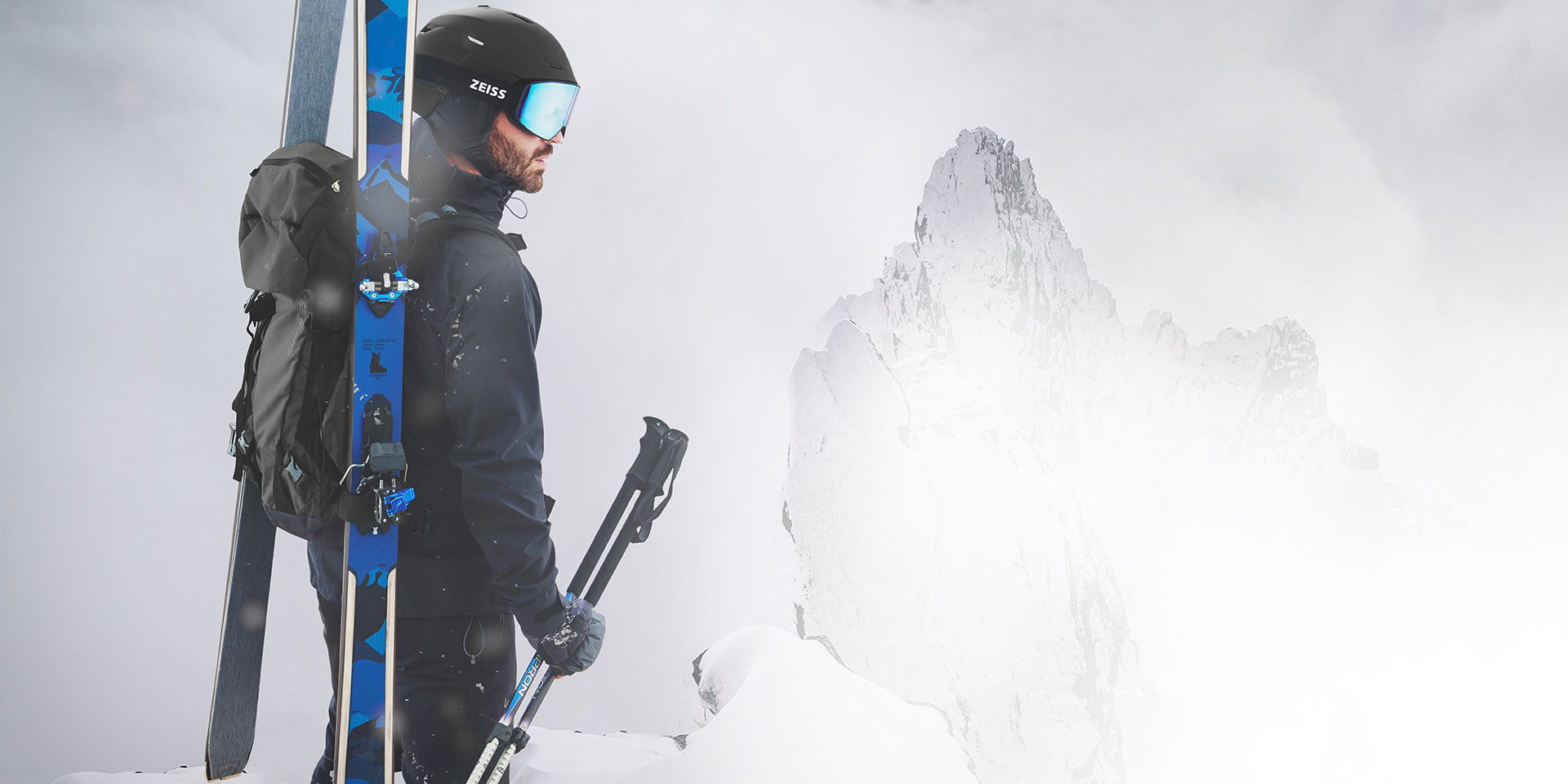 Un hombre luce un casco negro con máscaras de esquí ZEISS azules espejadas, lleva esquís y una mochila sobre la espalda y sujeta los bastones de esquí con la mano derecha. Está de pie en la cima de una montaña.