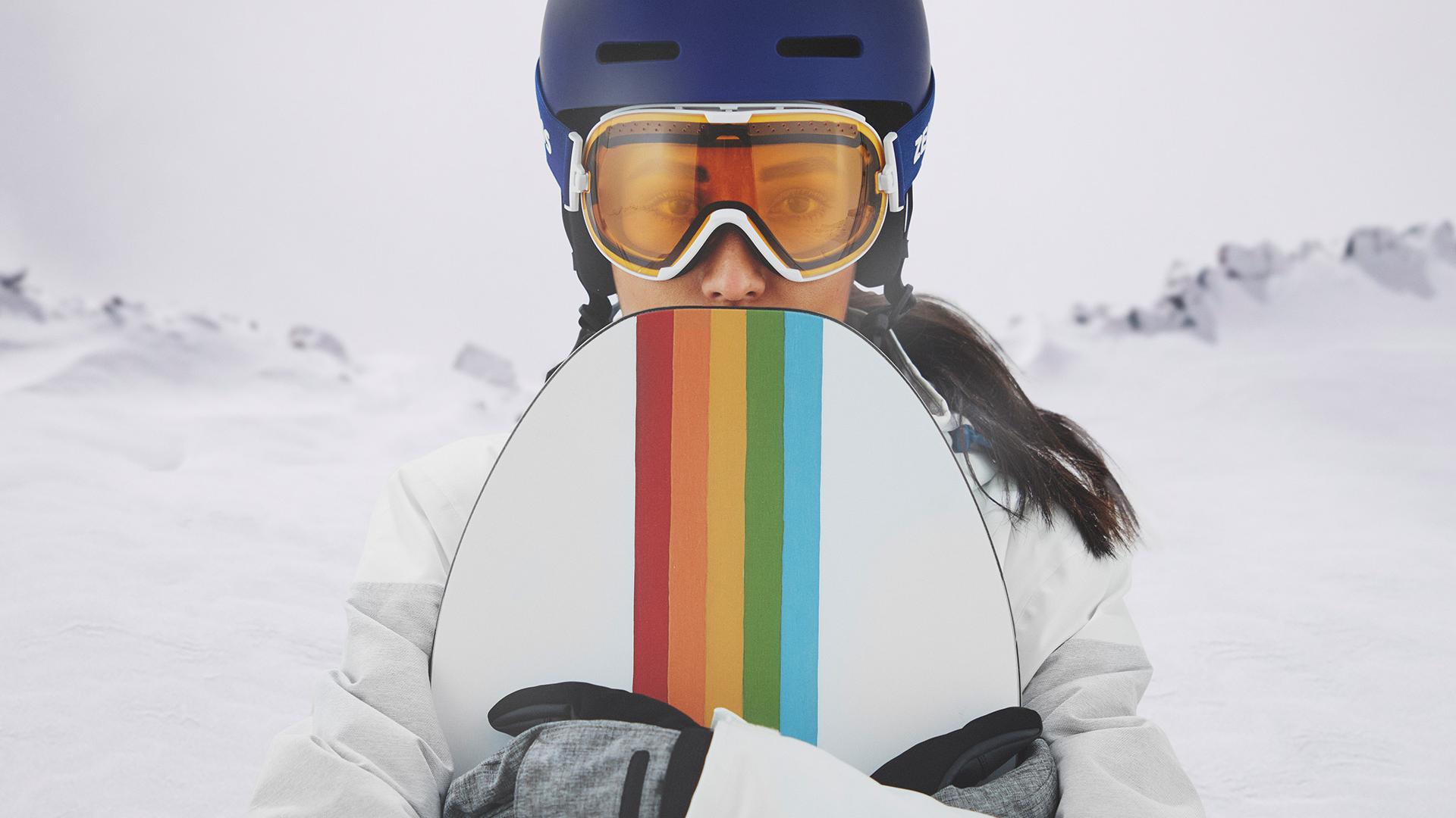 Una joven mujer que usa máscaras de nieve con tecnología ZEISS Interchangeable y sujeta una tabla de snowboard.